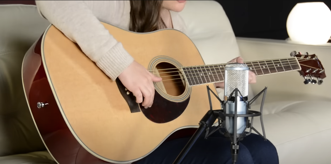 Photo of a girl playing a guitar while seated on a sofa, with a microphone focused on the guitar